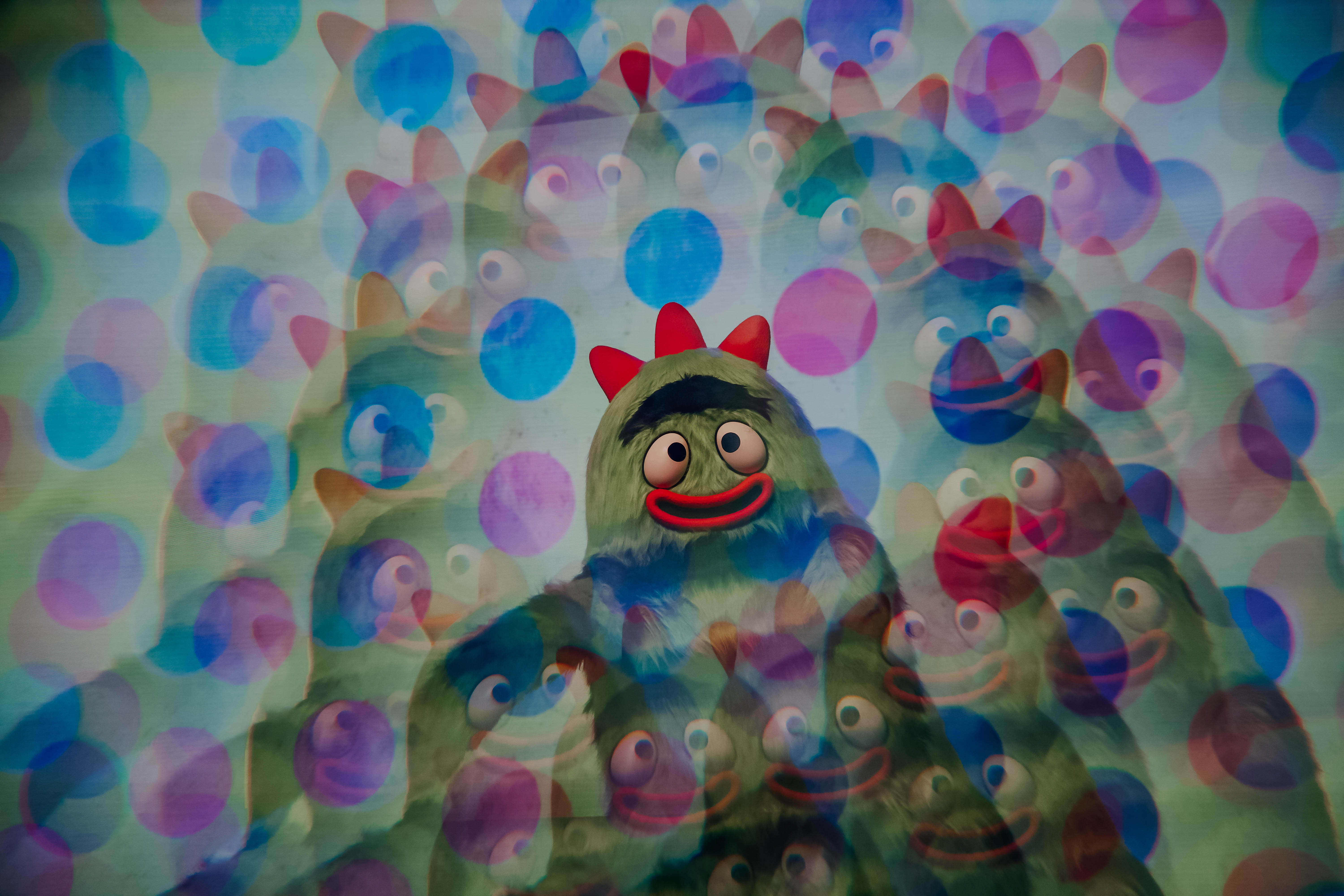 A colorful, whimsical character from a children's show, surrounded by a playful, blurred backdrop of similar characters and vibrant circles.