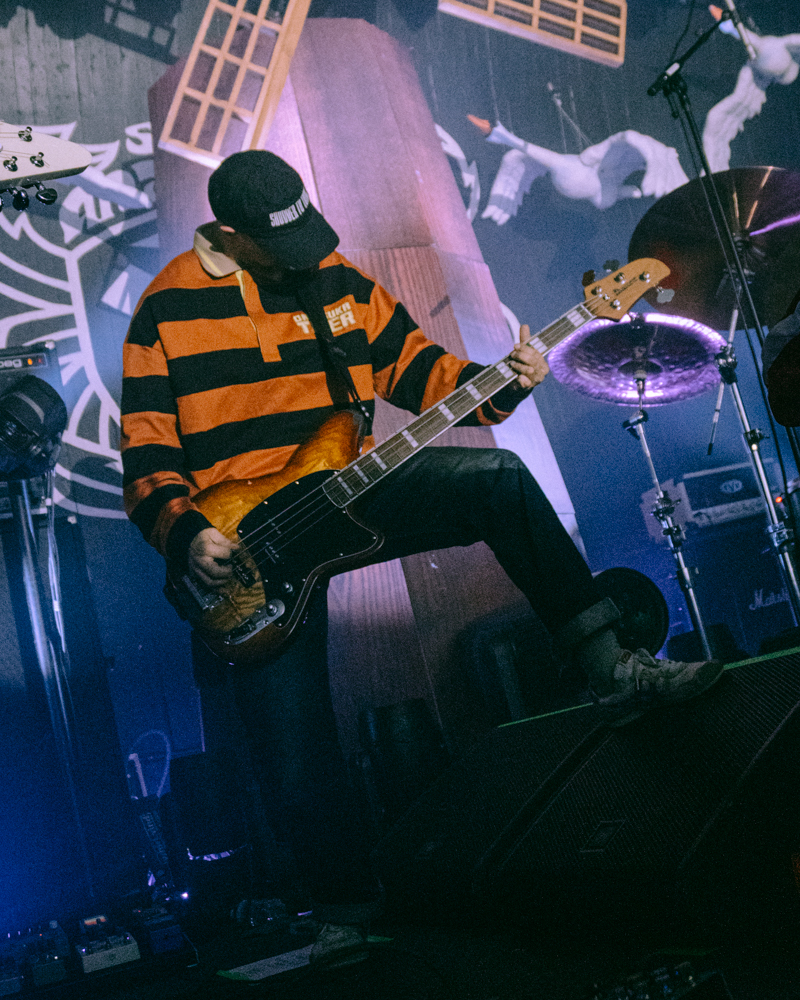 A musician playing bass guitar on stage, wearing a striped orange and black jersey and a baseball cap, with vibrant lighting and musical equipment in the background.