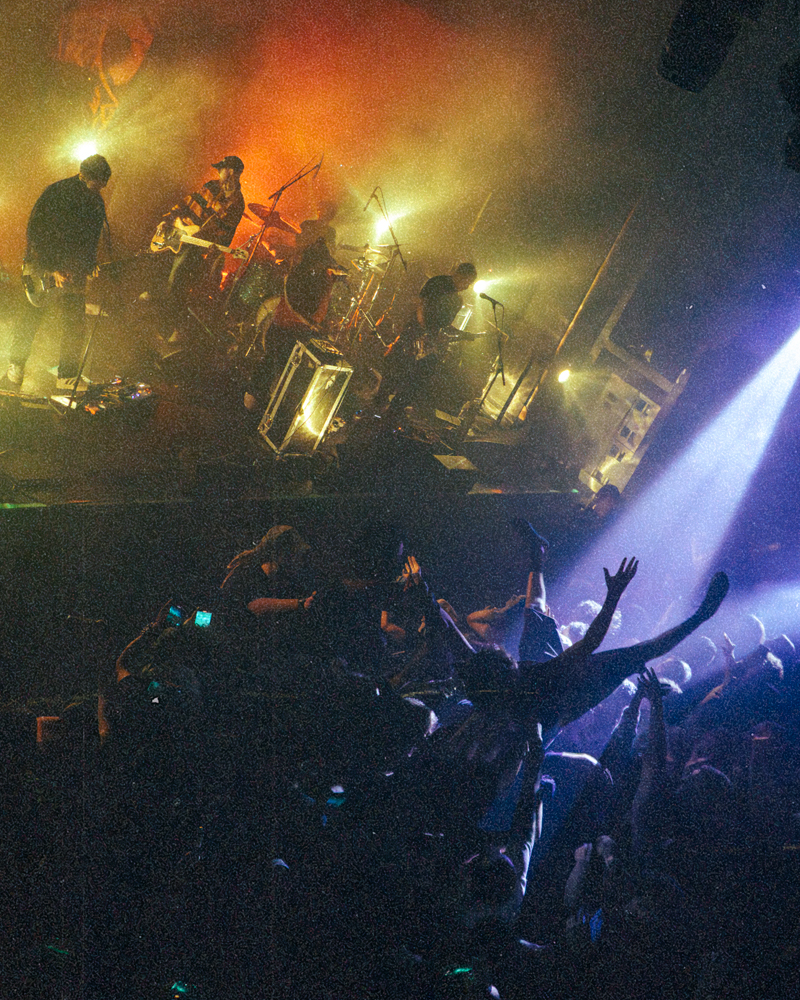 A live concert scene featuring the band Fleshwater performing on stage, with colorful lights and a crowd actively engaged in crowd-surfing and moshing.