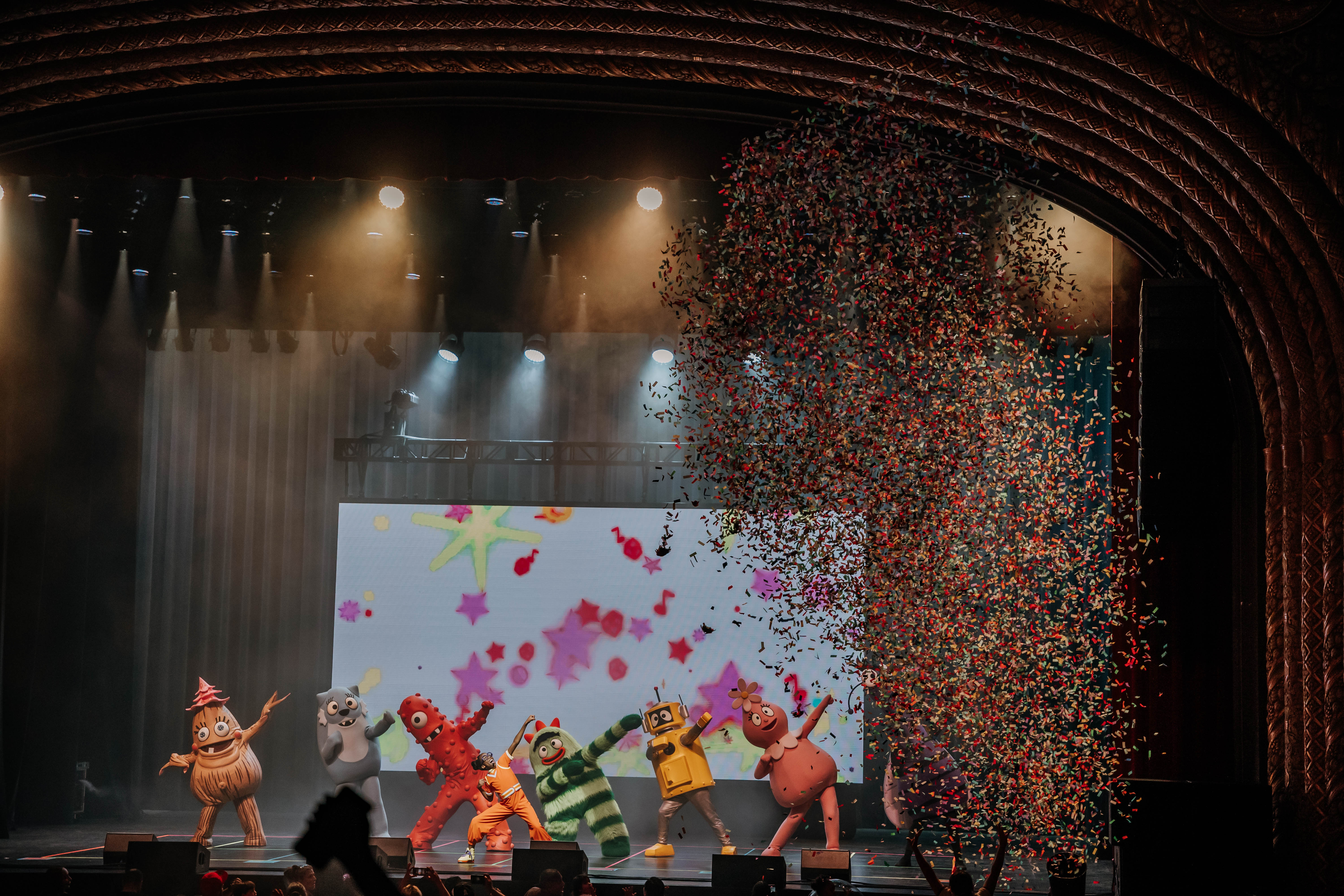 Colorful confetti falls from above as characters from Yo Gabba Gabba! perform on stage, surrounded by bright lights and animated backgrounds.