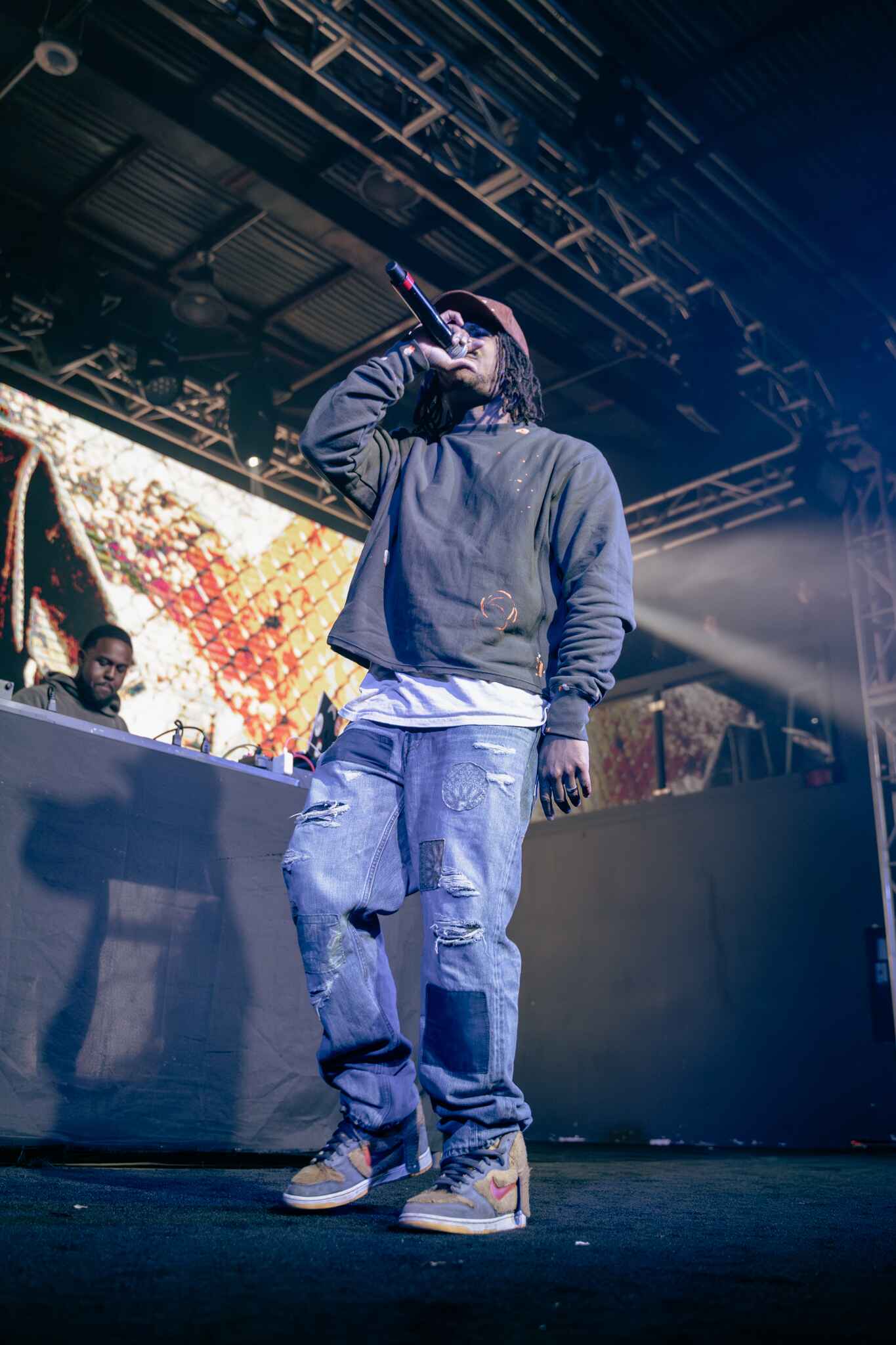 A performer holding a microphone on stage, wearing a black sweatshirt and distressed jeans, with a DJ in the background.