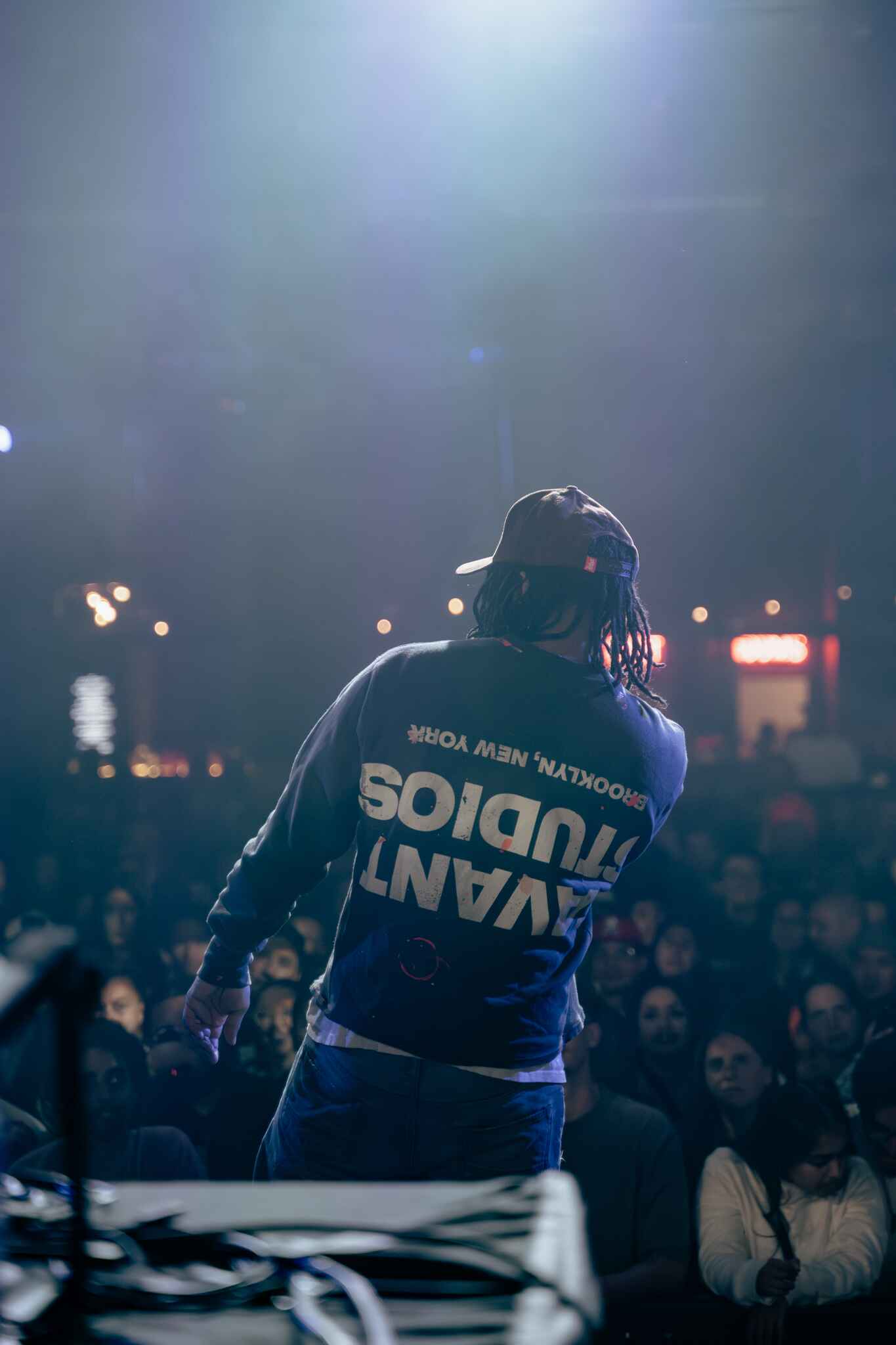 A performer on stage with their back to the camera, wearing a dark sweatshirt with 'Brooklyn, New York' printed on it. The crowd is visible in front, showing enthusiasm as they watch the performance.