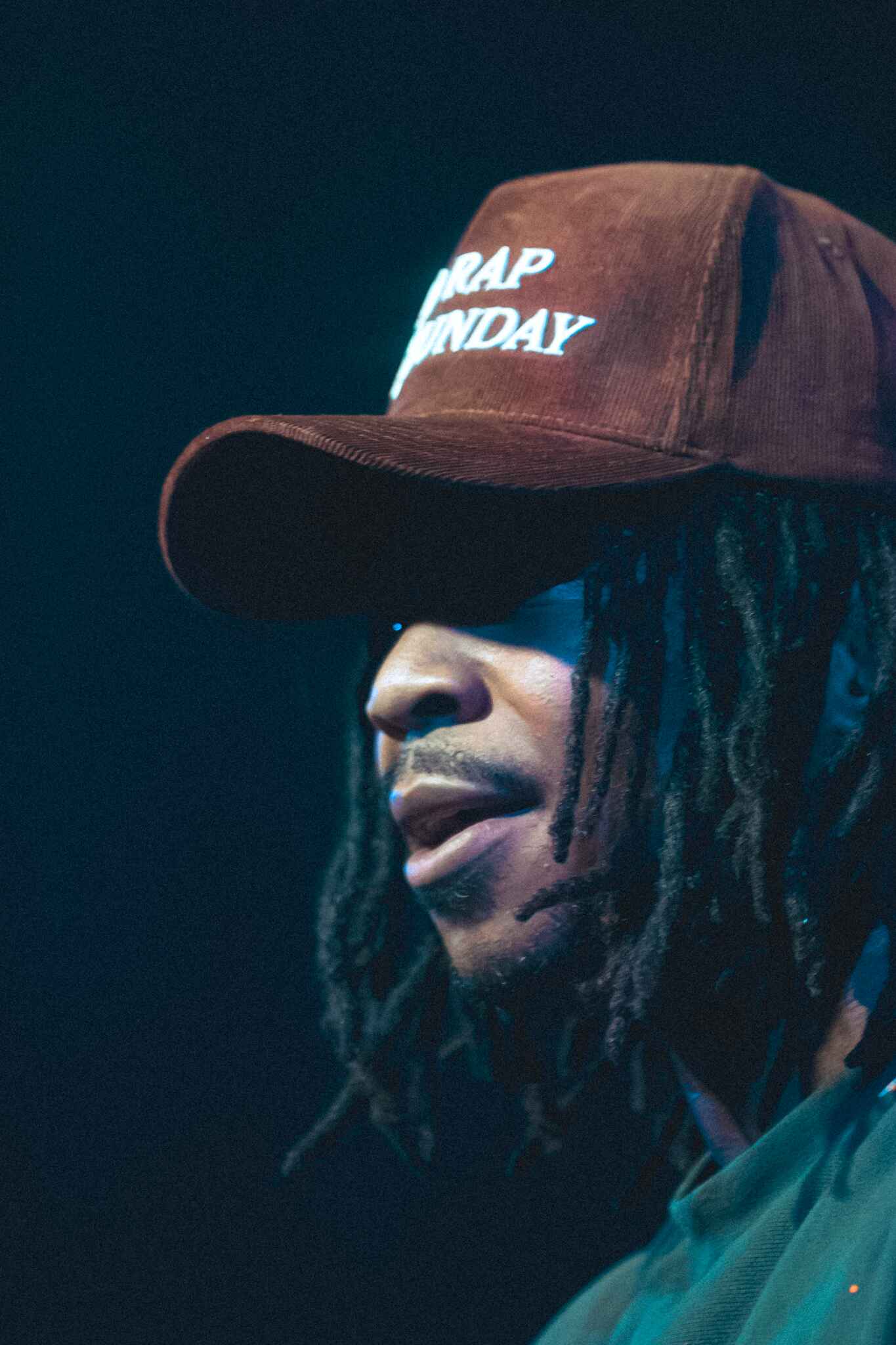 Close-up of a male performer wearing a brown cap with 'No Rap Sunday' written on it, with a focused expression.