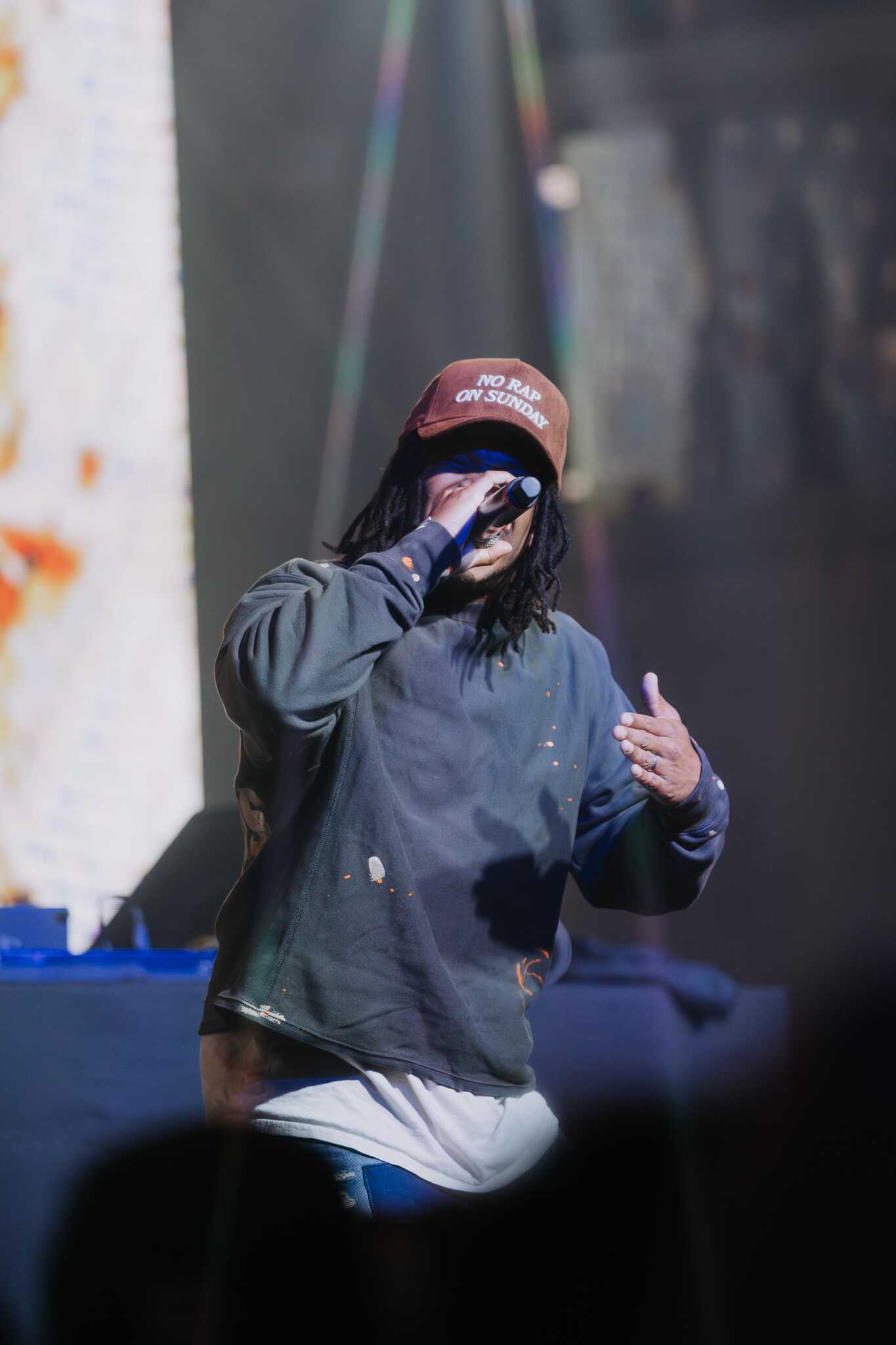 A musician performing on stage, wearing a brown hat with the text 'NO RAP ON SUNDAY' and a gray sweatshirt. The background features colorful lighting and screens displaying visuals.