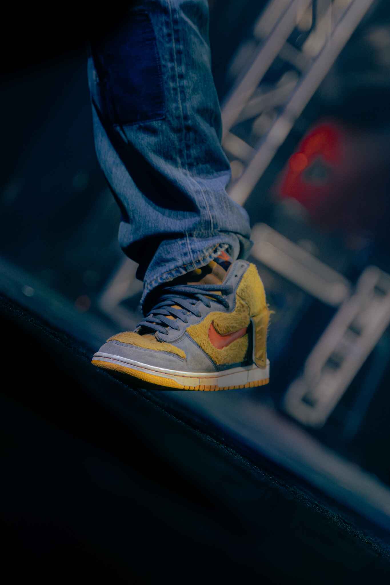 Close-up of a stylish sneaker worn by a performer, featuring a mix of yellow, gray, and orange colors against a blurred concert background.