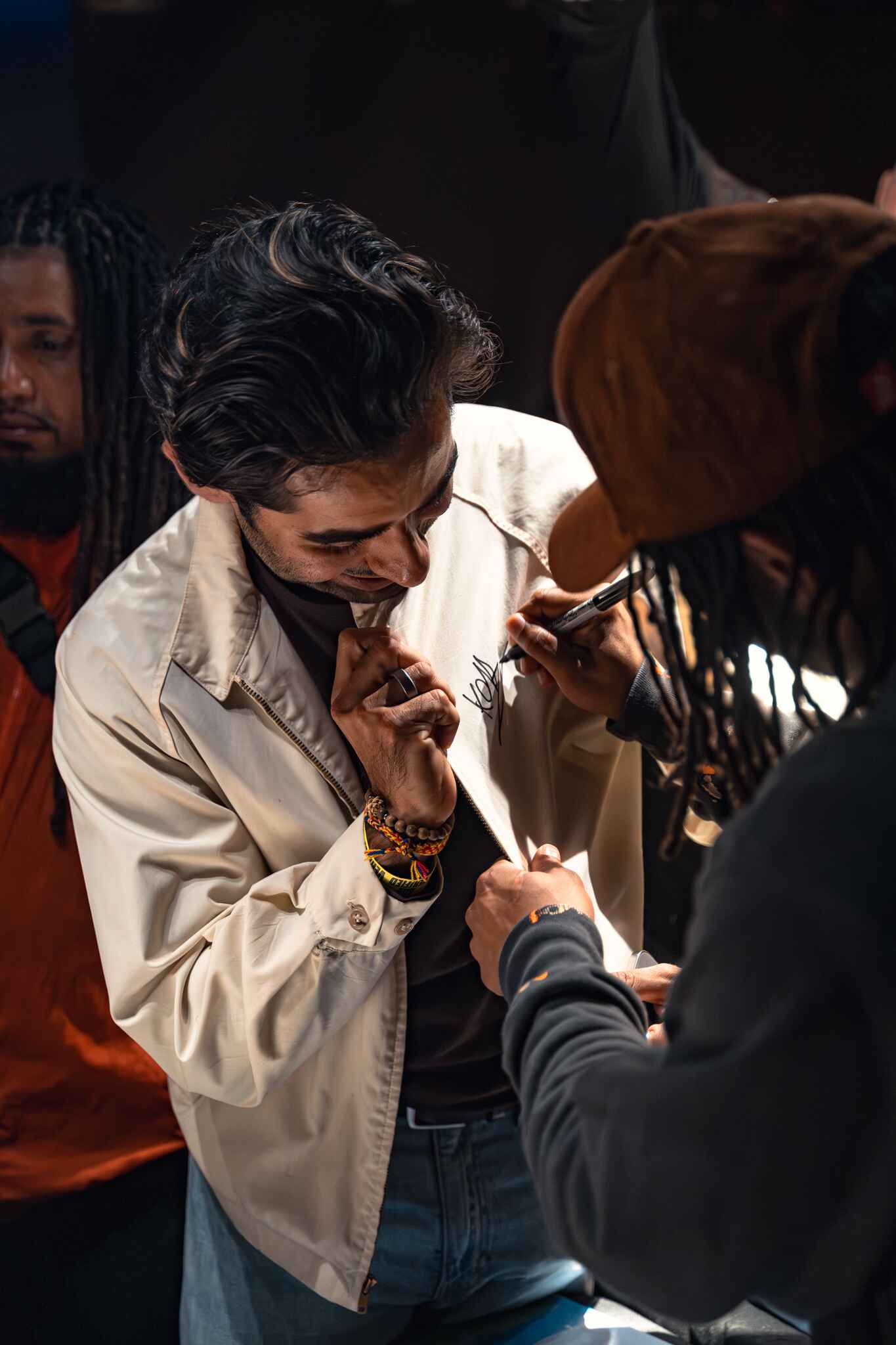 A person with dark hair is smiling while getting an autograph signed on their jacket by another person. In the background, another individual with dreadlocks is visible.