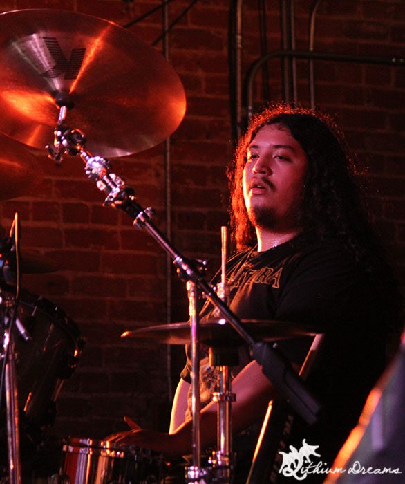 A drummer performing on stage with long hair and wearing a band t-shirt, captured in a dimly lit venue with a brick wall background.