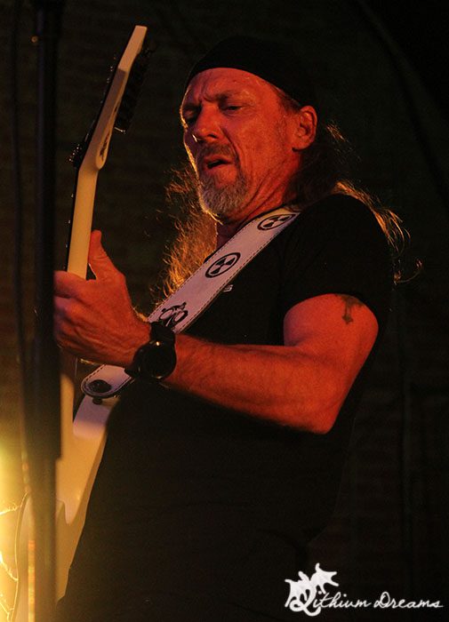 Close-up of a guitarist performing on stage, wearing a black t-shirt and a headband, playing a white electric guitar with focused expression.