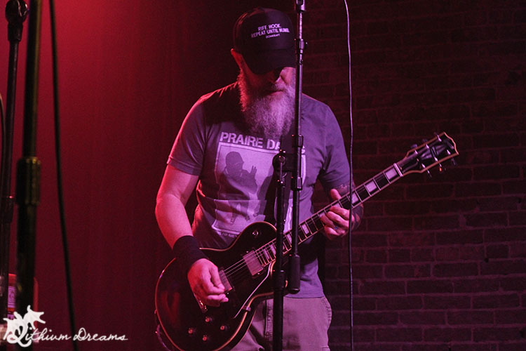 A guitarist with a beard playing an electric guitar on stage, wearing a 'Prairie Dog' t-shirt and a cap, with vibrant stage lighting in the background.