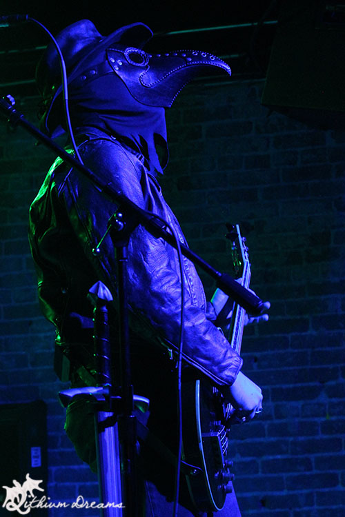 A musician wearing a plague doctor mask and leather outfit plays an electric guitar on stage, illuminated by blue and green lighting.