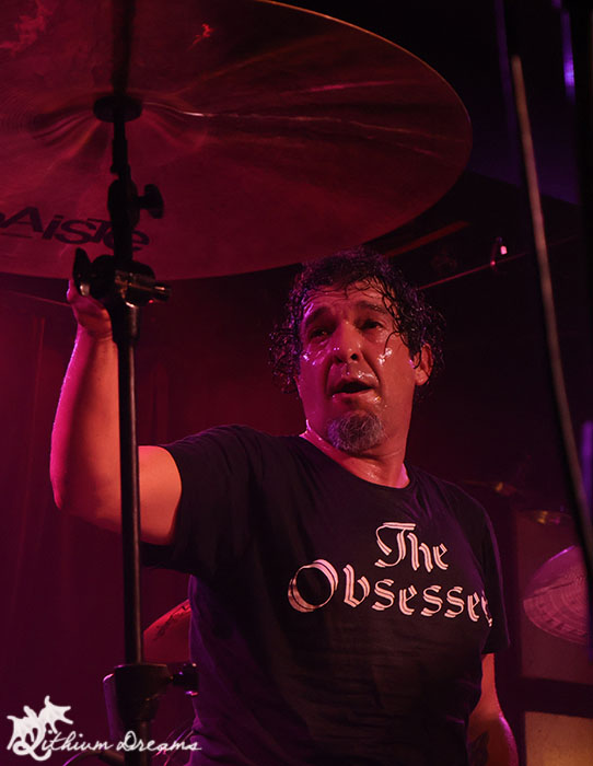 Close-up of a drummer performing on stage, wearing a black shirt with the text 'The Obsesser'. The drummer appears intense and focused, with sweat visible on his face under colorful stage lighting.