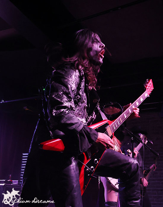 A guitarist performing passionately on stage, wearing a black patterned coat and making dynamic gestures while playing a red electric guitar.