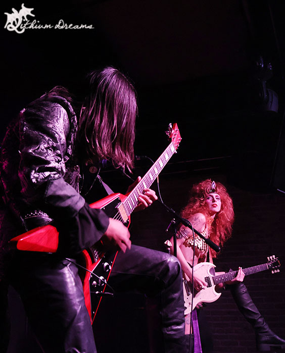 A guitarist performing on stage with a dynamic pose, while a female vocalist stands nearby, dressed in an elaborate outfit with long, curly hair, all set against a dark ambiance.