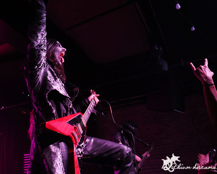 A performer in a dramatic outfit plays an electric guitar on stage, passionately singing with arms raised, while animated audience members show their excitement.