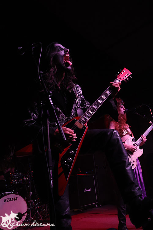 A guitarist performing passionately on stage, with vibrant stage lights illuminating the scene, while a vocalist plays a bass guitar nearby. The drummer is visible in the background.