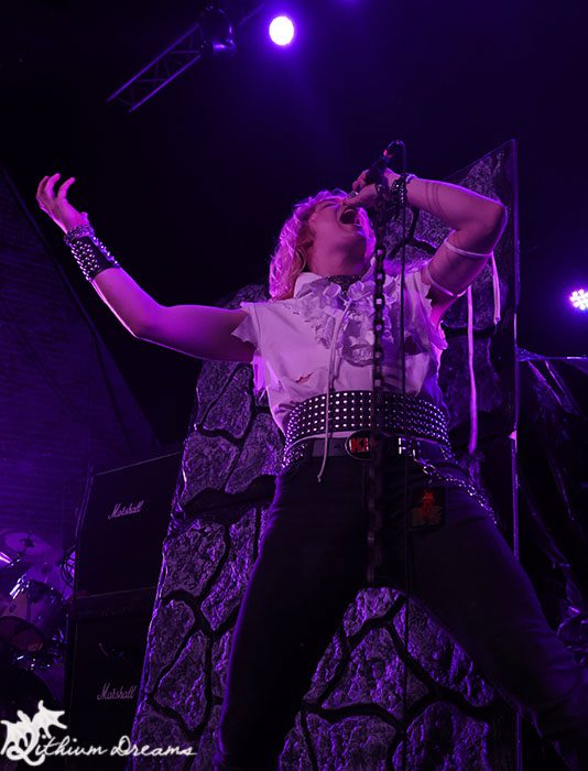 A thrash metal vocalist passionately performing on stage with arms raised, sporting a vintage rock outfit and a belt with spikes, against a backdrop featuring amplifiers and colorful stage lights.