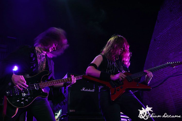 Two male musicians performing on stage, one playing a bass guitar and the other playing an electric guitar, with energetic stage presence and dramatic lighting.