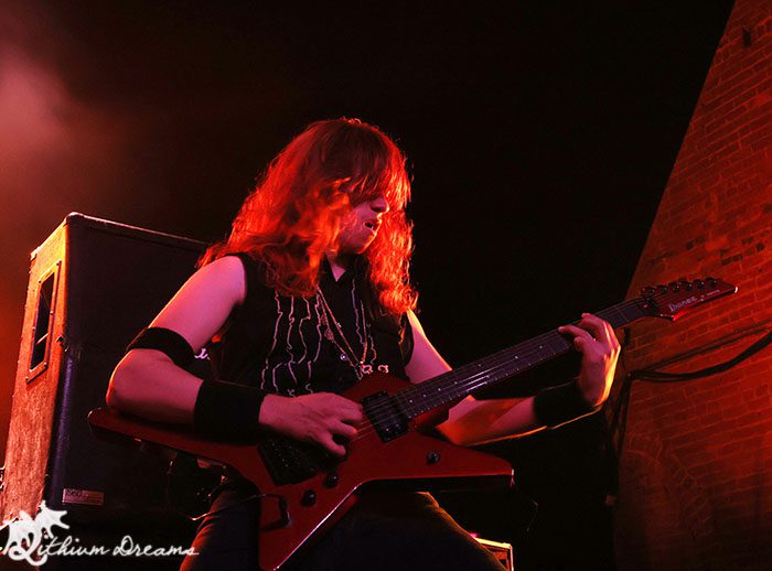 A guitarist with long red hair performing on stage, wearing a black sleeveless shirt and playing a red electric guitar, with dramatic stage lighting in the background.