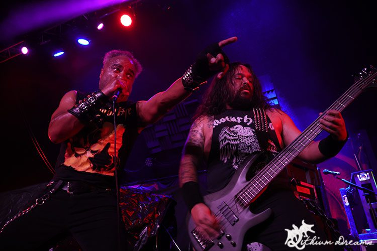A dynamic scene capturing two male musicians performing on stage during a metal concert. The vocalist, with distinct hair and attire, engages the audience energetically, while the bassist plays intensely, both surrounded by vibrant stage lights.
