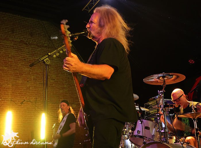 A live performance featuring a bassist singing into a microphone, with a drummer in the background and another musician playing guitar.