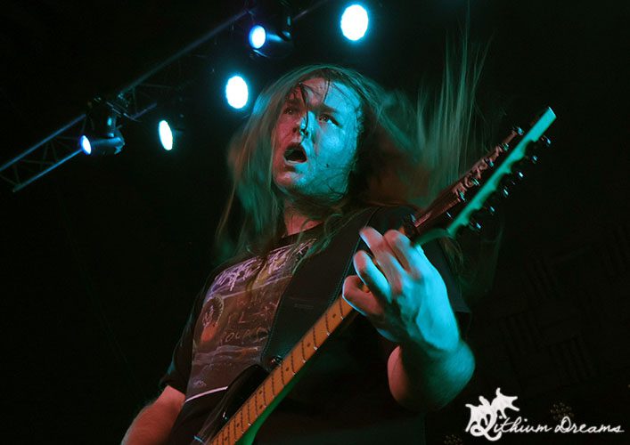 A guitarist with long hair passionately playing on stage under bright blue lights.