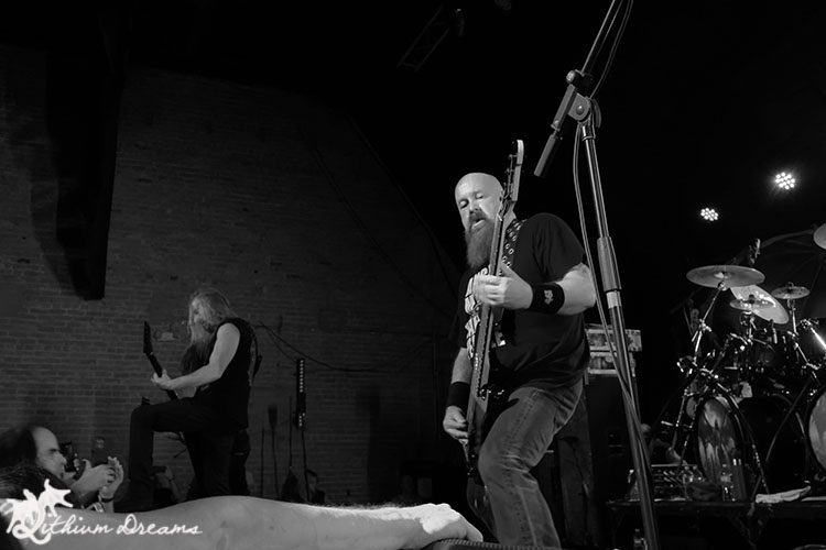 A black and white image of a live thrash metal performance, featuring a band on stage with one musician playing guitar and another in the background, emphasizing an energetic and intense atmosphere.