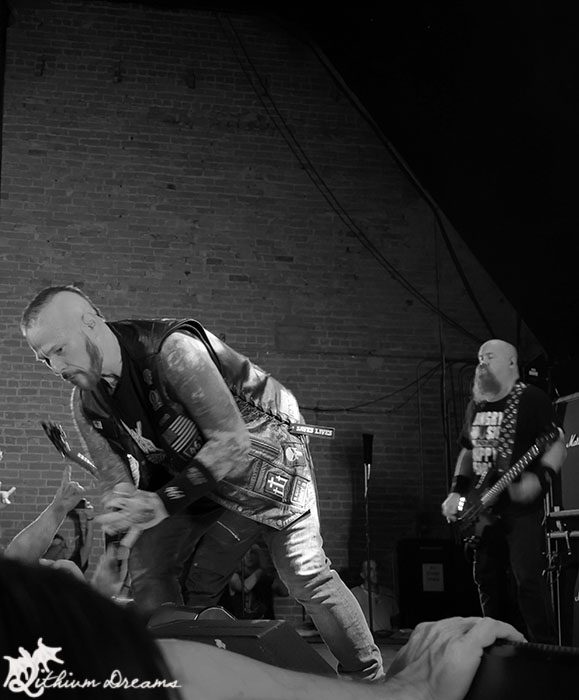 A live performance at the Nile Theater featuring a thrash metal band, with a lead vocalist engaged with the audience, while a guitarist plays in the background. The venue showcases brick wall architecture.