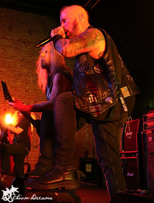 Two male musicians performing on stage, one singing into a microphone while the other plays electric guitar, with a brick wall and sound equipment in the background, illuminated by stage lighting.