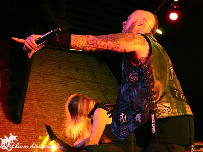 A live performance shot of a heavy metal band on stage, featuring a vocalist with tattoos and a sleeveless vest, energetically engaging the audience. The background showcases dramatic lighting and brick walls, creating a vibrant concert atmosphere.