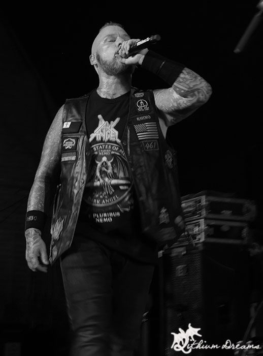A vocalist passionately performing on stage during a metal concert, wearing a black vest with band patches and a t-shirt, microphone in hand.