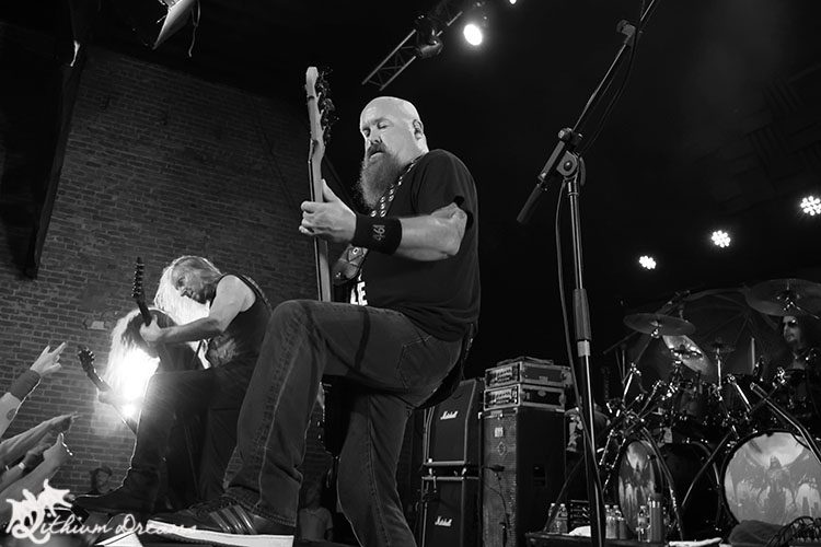 A black and white image of a live metal band performance, featuring two guitarists and a bassist on stage, with the audience's hands raised in front.