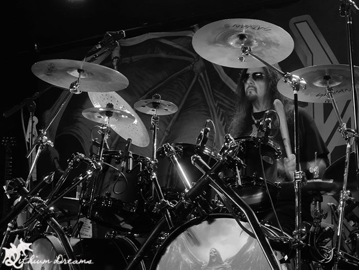 A drummer performing on stage with sunglasses, surrounded by a drum set and cymbals, set against a backdrop with a graphic design.