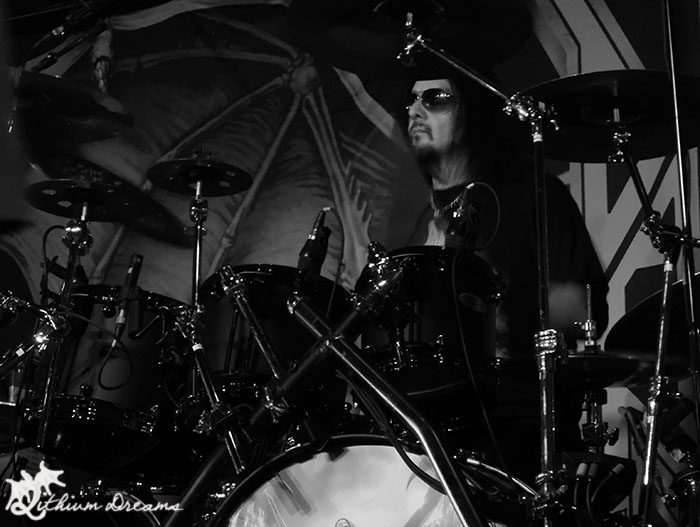 A drummer performing on stage, wearing sunglasses, with a dark background featuring a skeletal illustration.