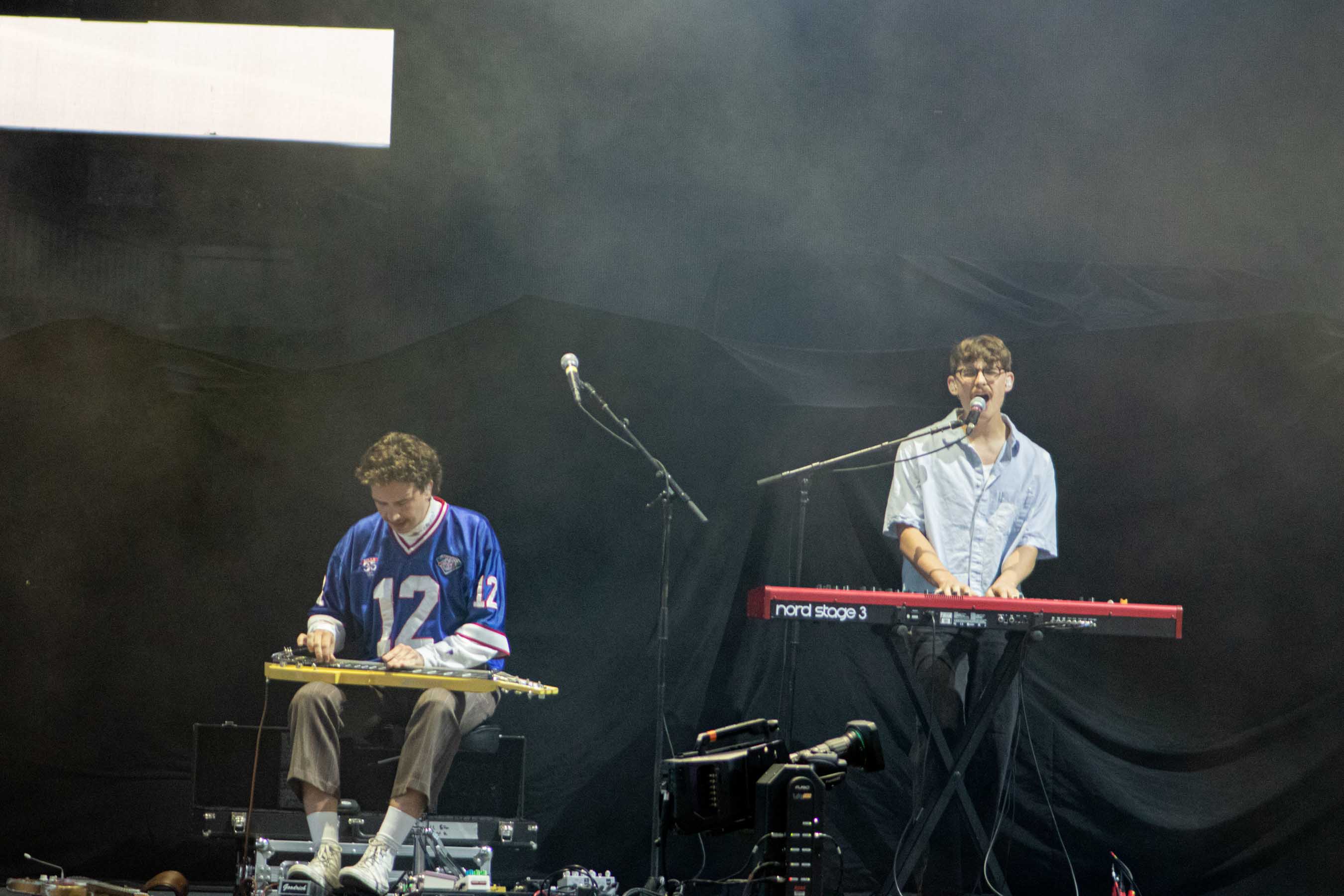 Two musicians performing on stage, one playing a steel guitar and the other singing into a microphone while playing a keyboard, with a dark backdrop and atmospheric lighting.