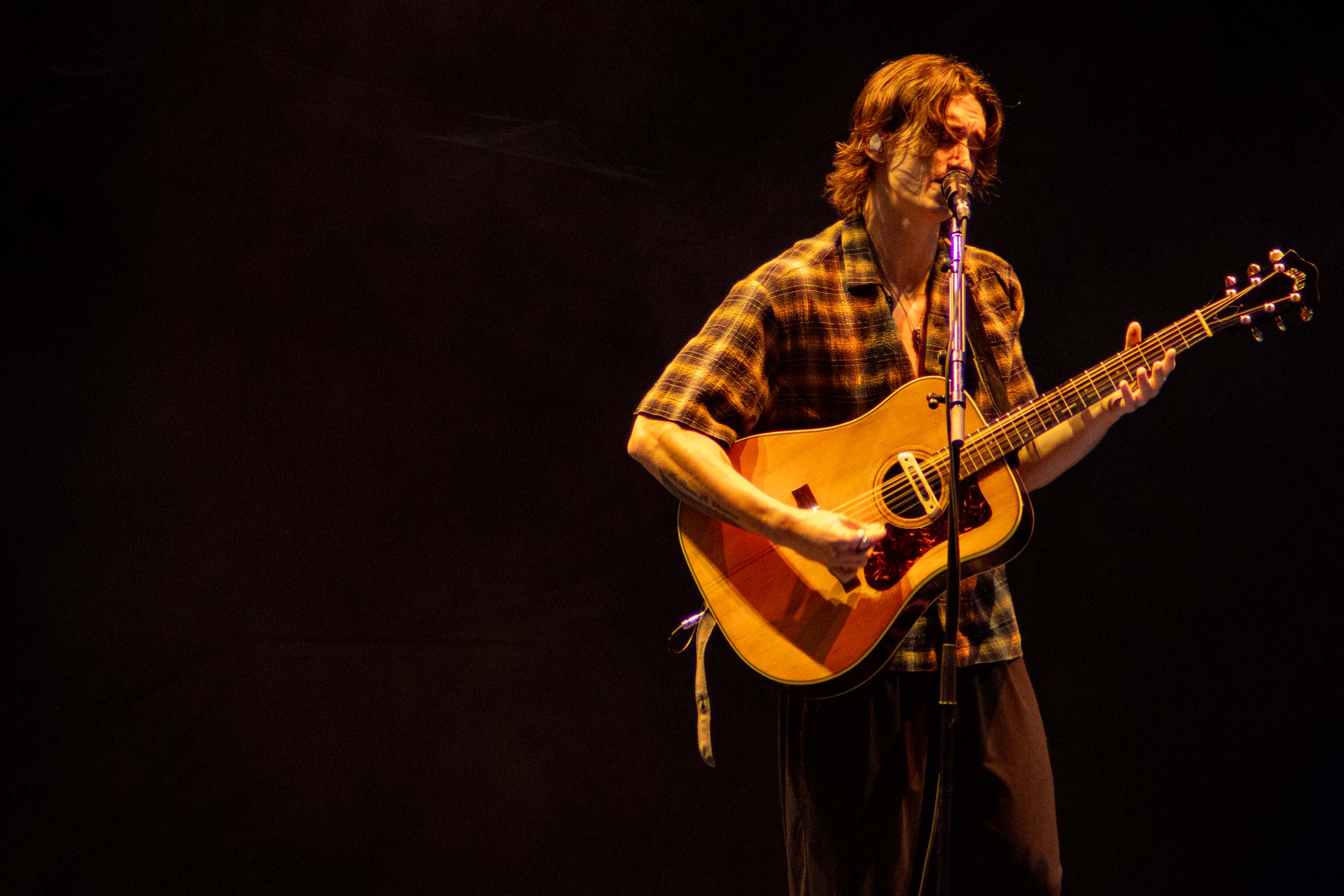 A musician performing on stage, playing an acoustic guitar while singing into a microphone, illuminated by stage lights.