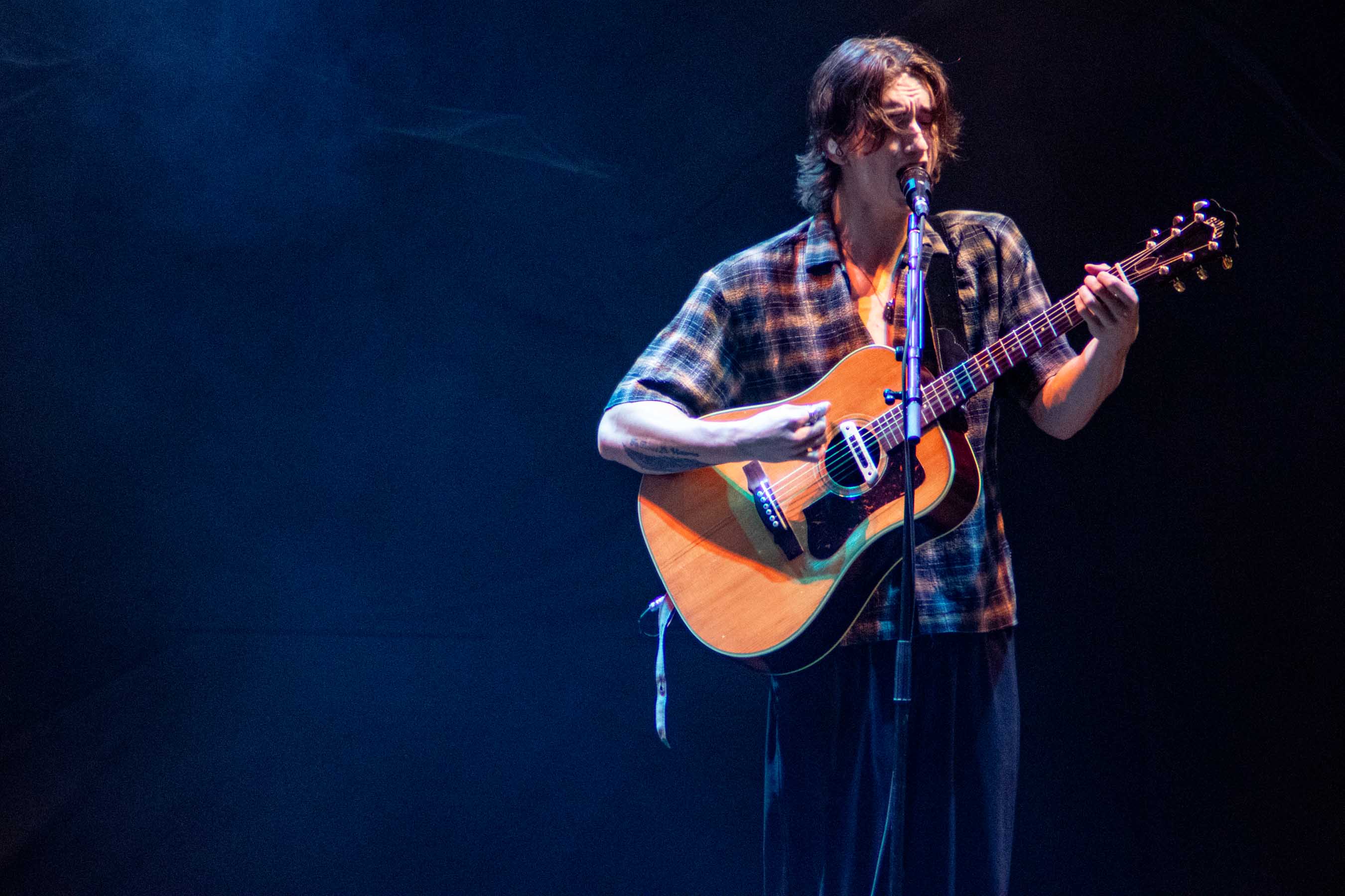 A musician performing on stage, passionately singing into a microphone while playing an acoustic guitar, dressed in a plaid shirt against a dramatic blue backdrop.