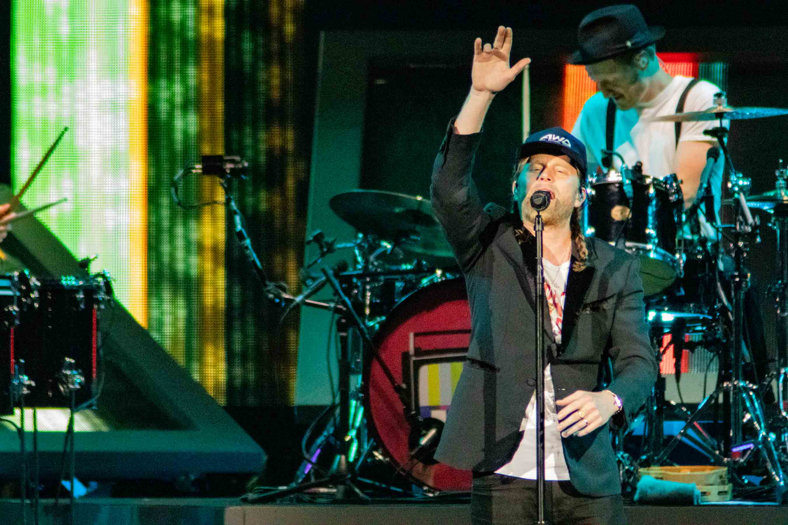 A male singer performing on stage with his hand raised, wearing a cap and a jacket, while a drummer is visible in the background.