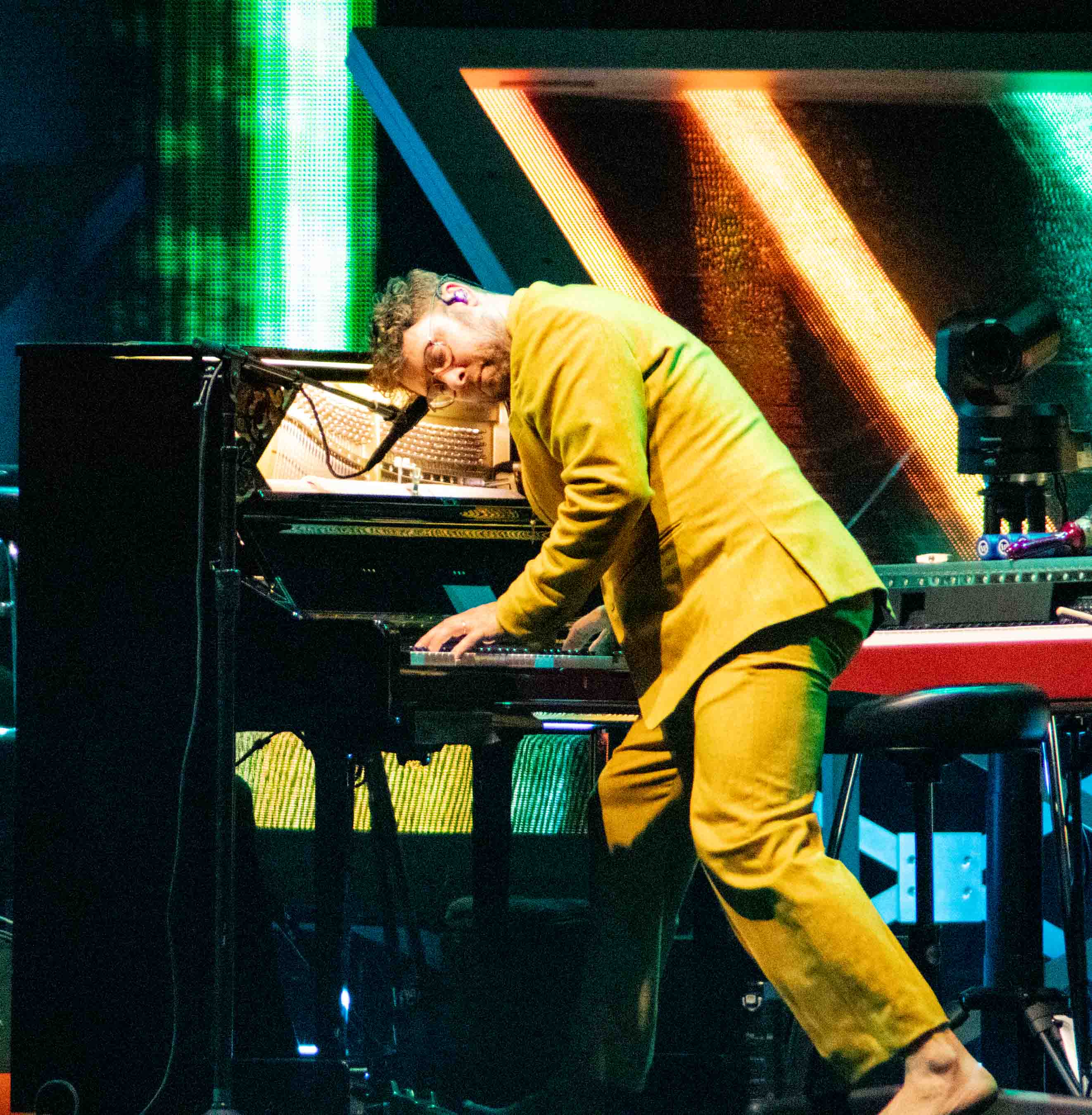 A musician in a bright yellow suit playing the piano during a live performance, surrounded by colorful stage lighting.