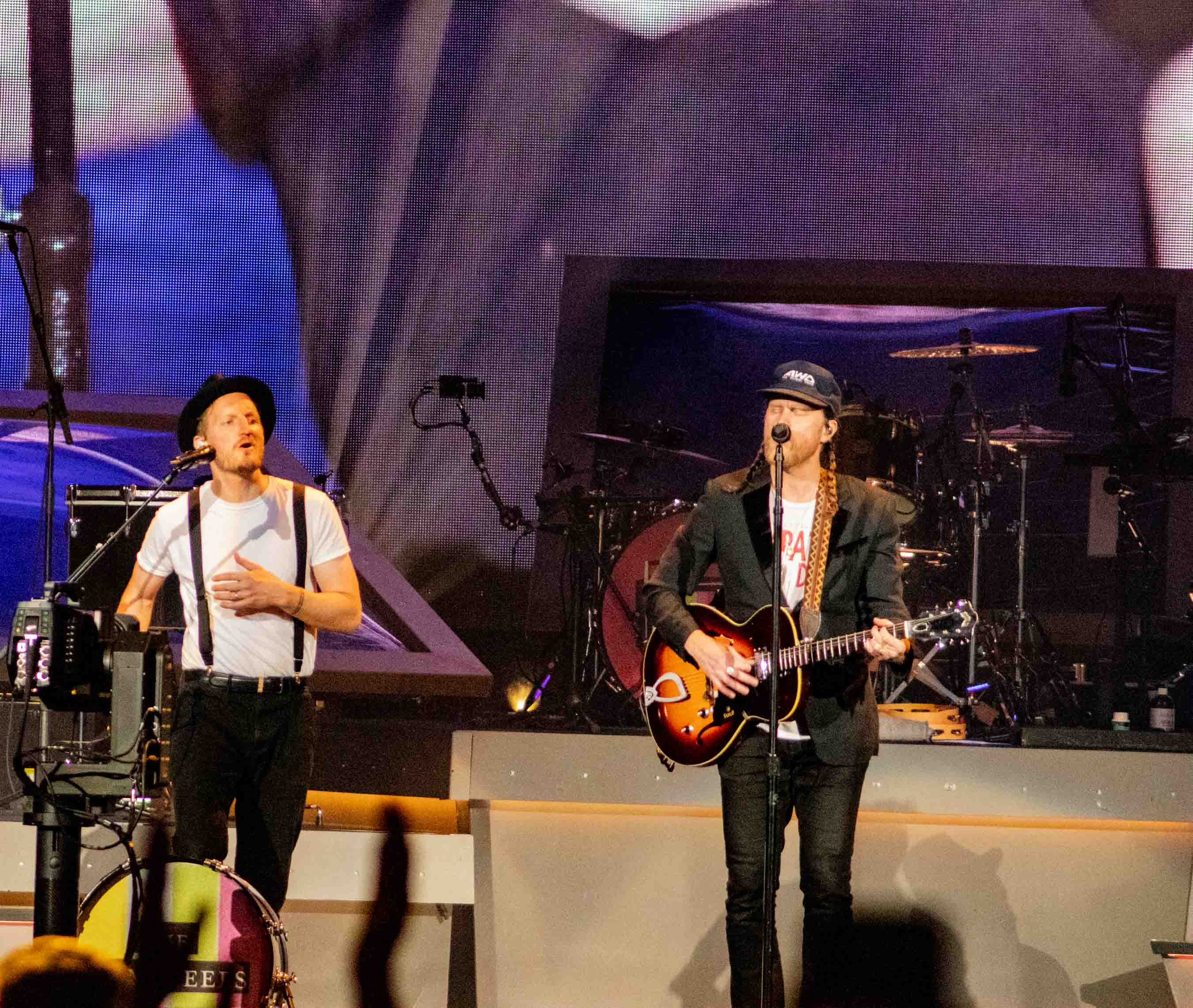 The Lumineers performing on stage, with one member playing guitar and singing while another provides vocals, showcasing a vibrant live concert atmosphere.