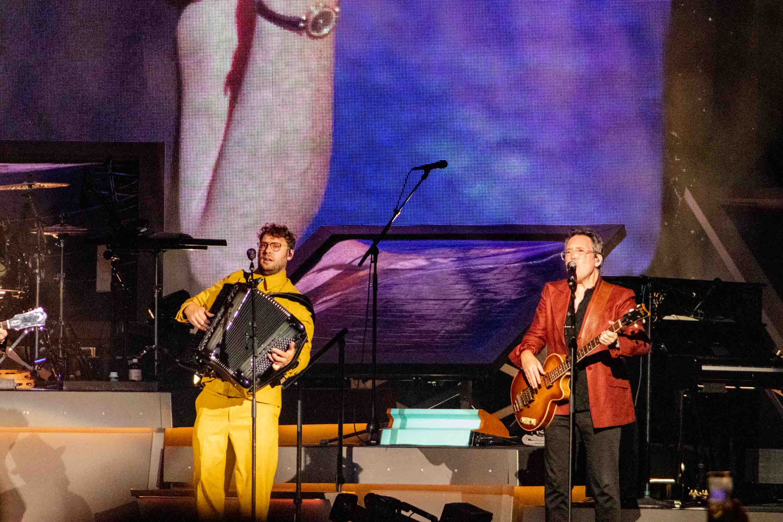 Two musicians performing on stage, one playing the accordion and the other with a guitar, set against a vibrant backdrop.