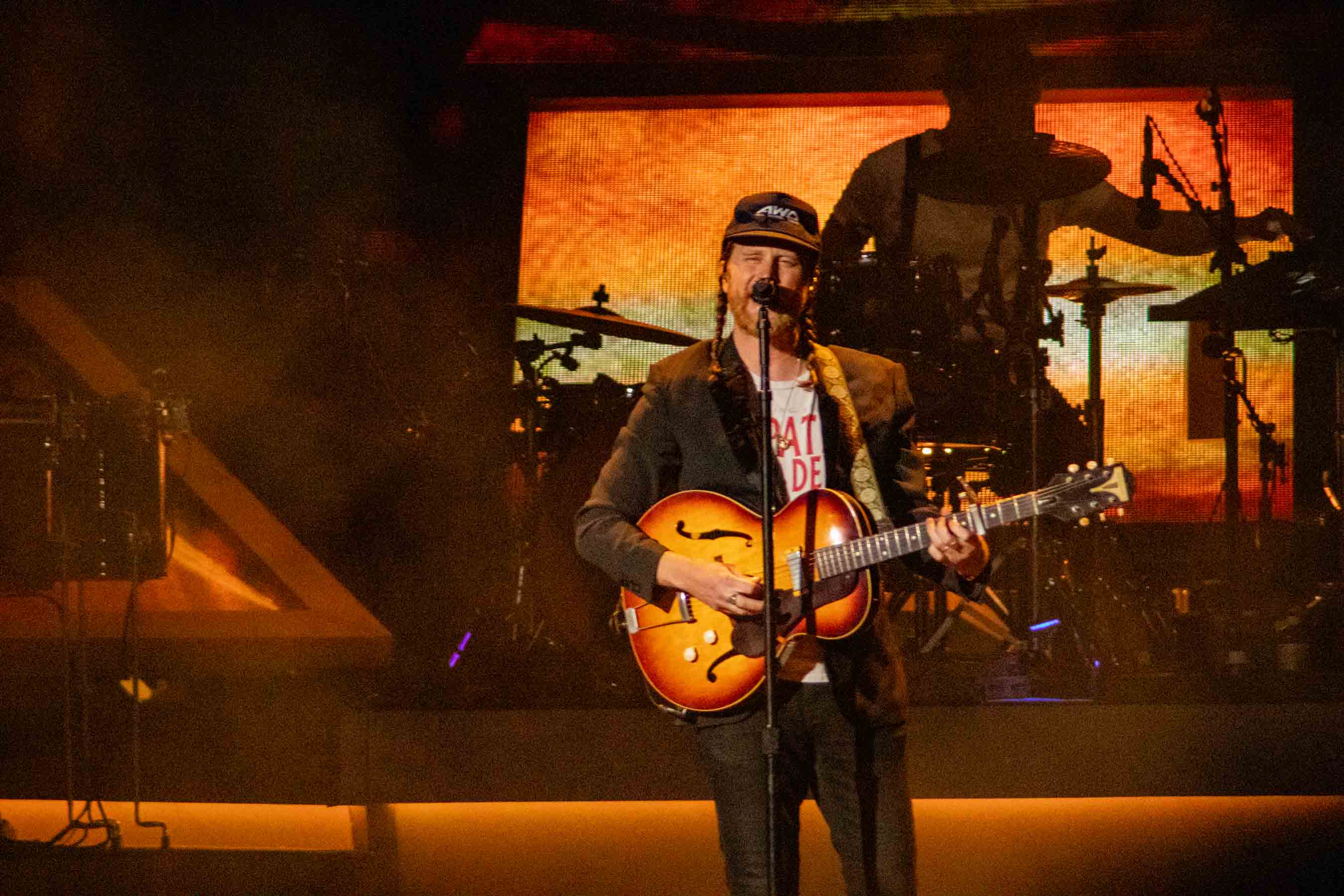 A musician performing on stage with an electric guitar, wearing a cap and a shirt, in front of a colorful backdrop with band instruments visible.