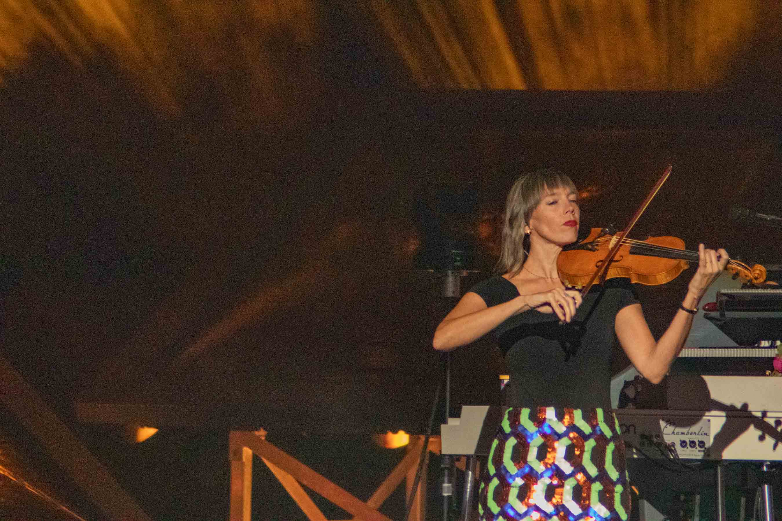 A female musician playing the violin on stage, wearing a black top and a colorful sequined skirt, with dramatic lighting in the background.