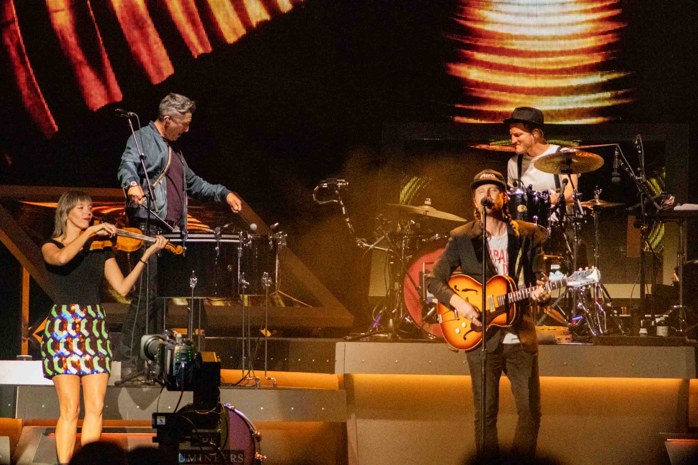 The Lumineers performing on stage, featuring a violinist, lead singer with an acoustic guitar, and a drummer, set against a vibrant light backdrop.