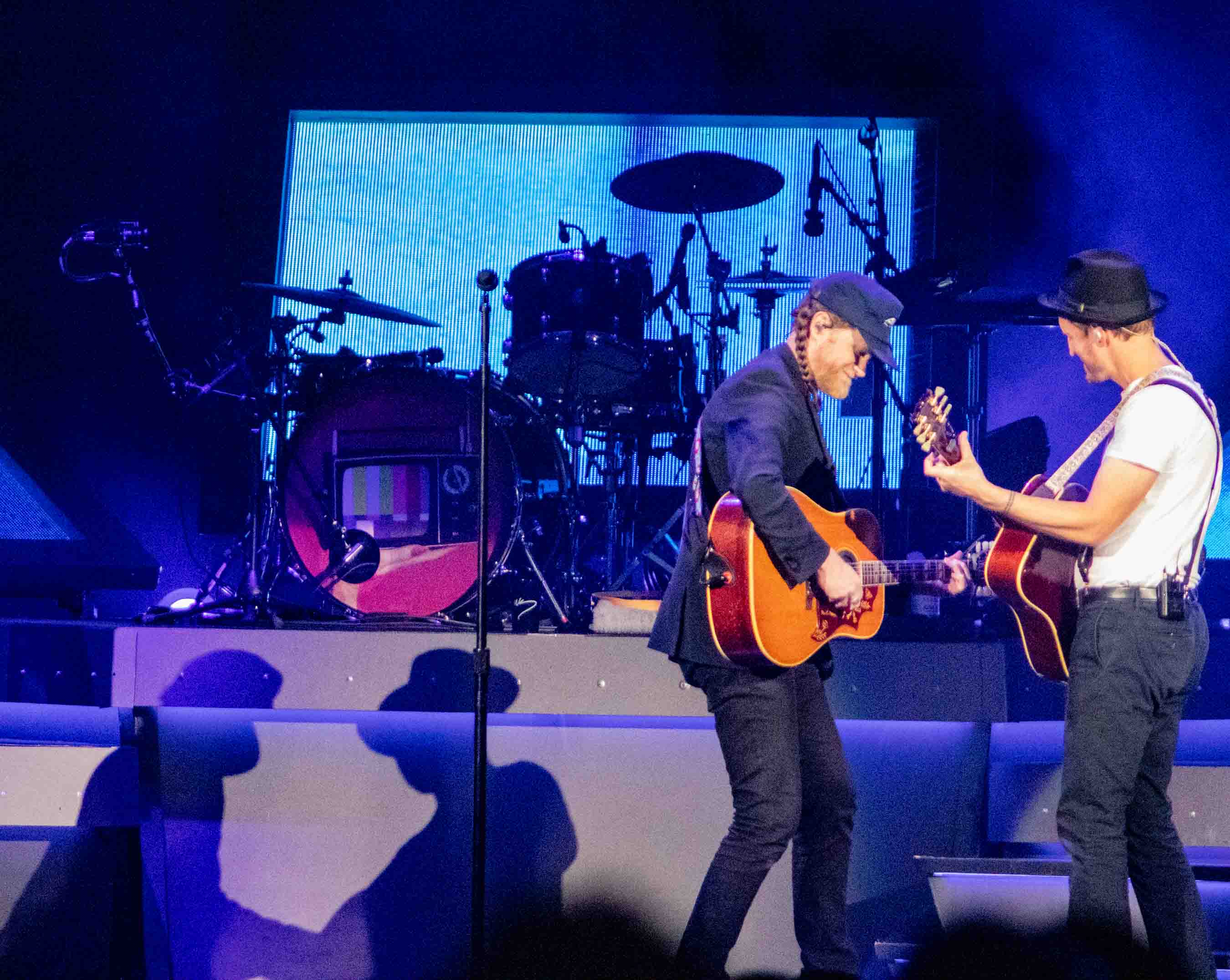 Two musicians performing on stage with guitars, set against a backdrop featuring drums and vibrant lighting.