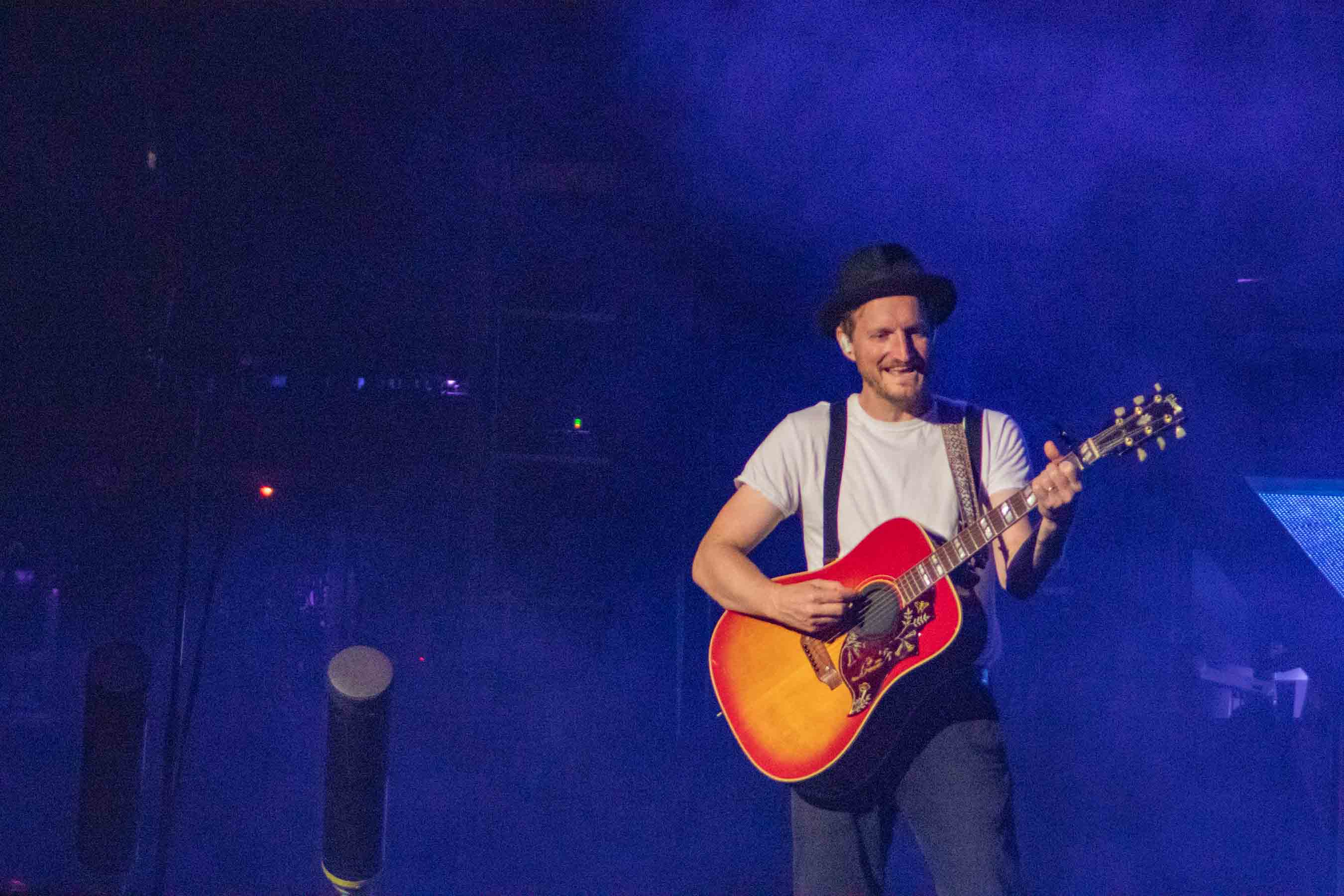 A musician performing on stage, smiling while playing an acoustic guitar, surrounded by a colorful stage lighting and a misty atmosphere.