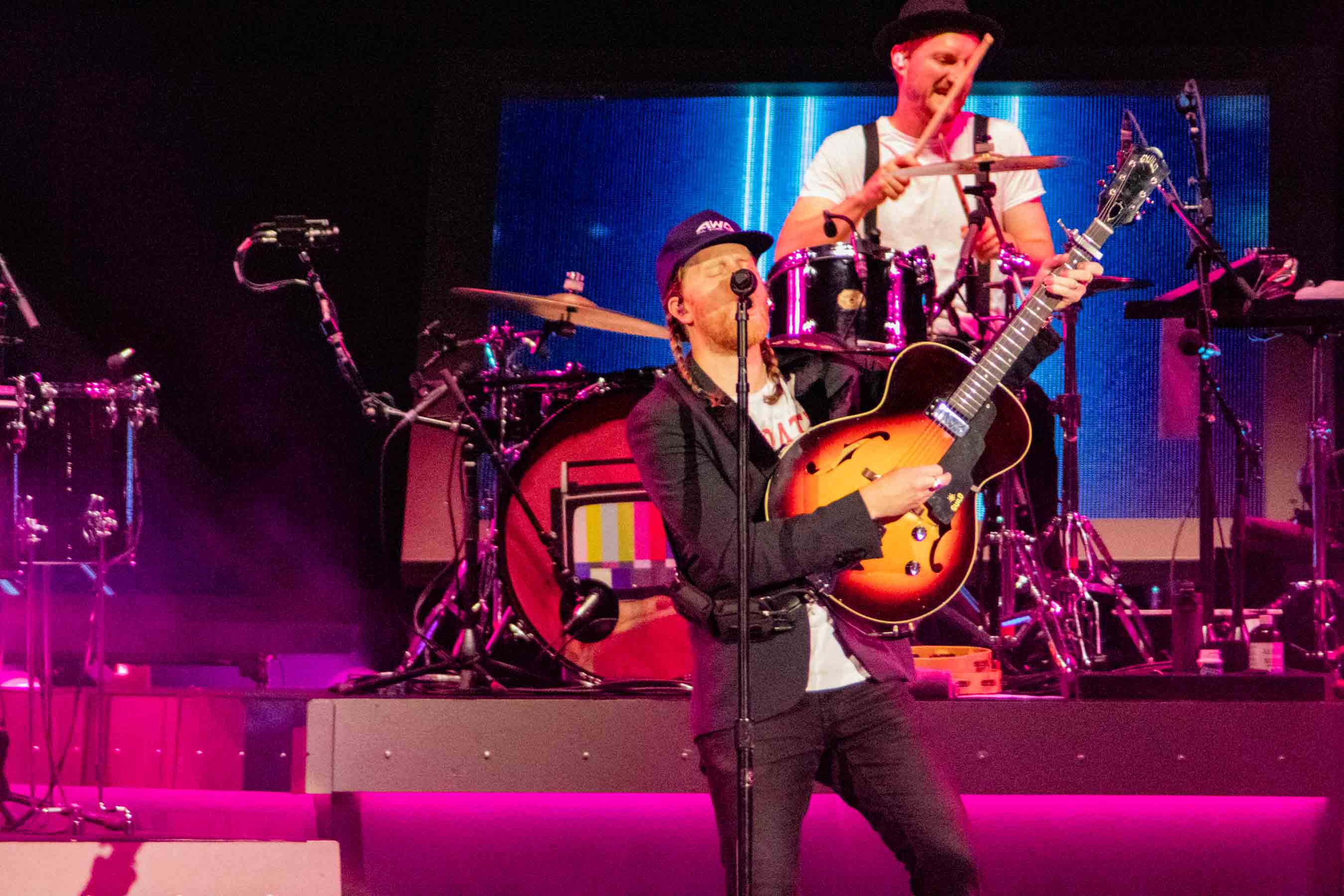 A performer energetically plays the guitar on stage, with a drummer in the background. The stage lights create a vibrant atmosphere.