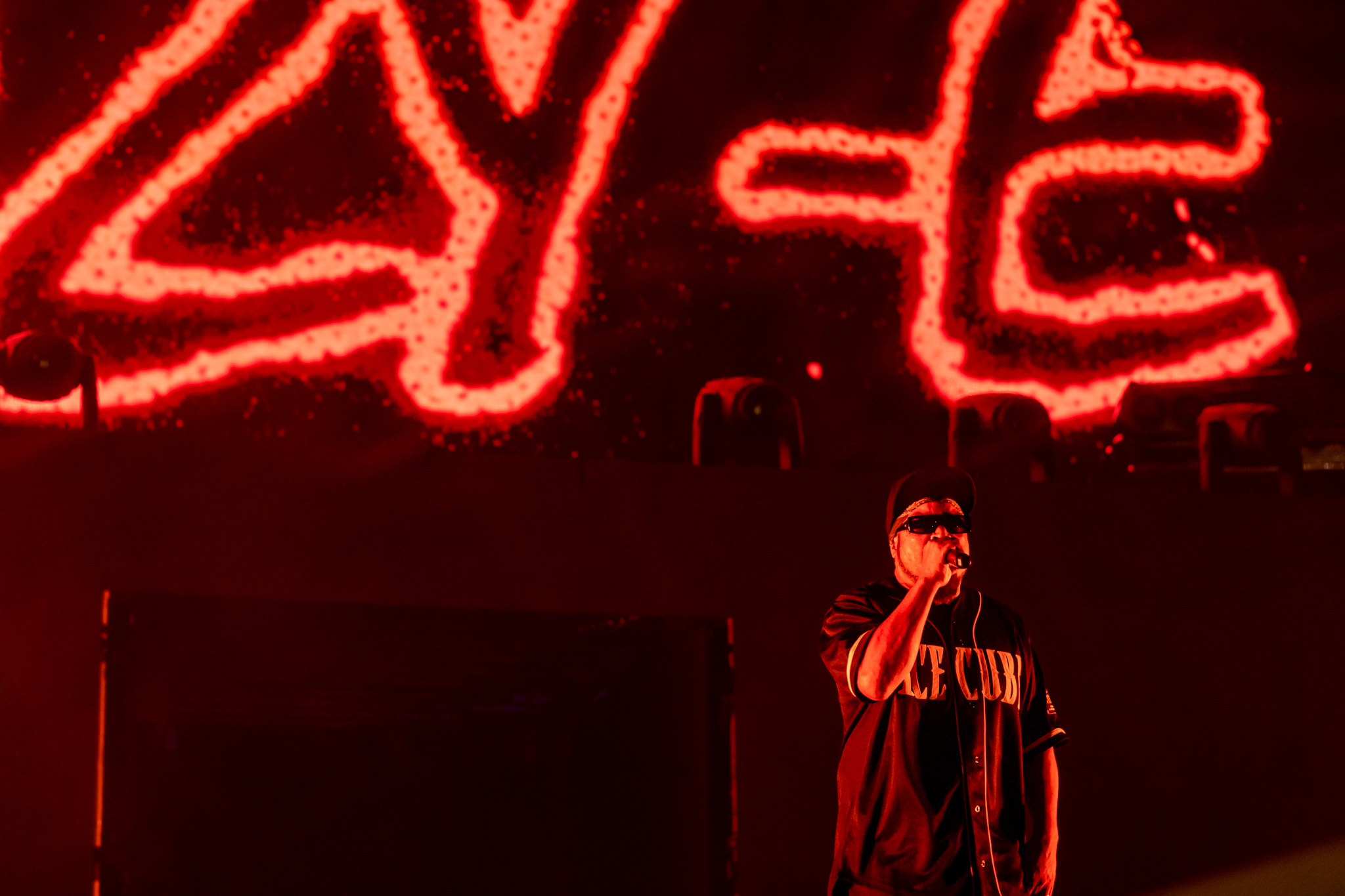 Ice Cube performing live on stage at Ball Arena with a dramatic red background displaying his name in lights.