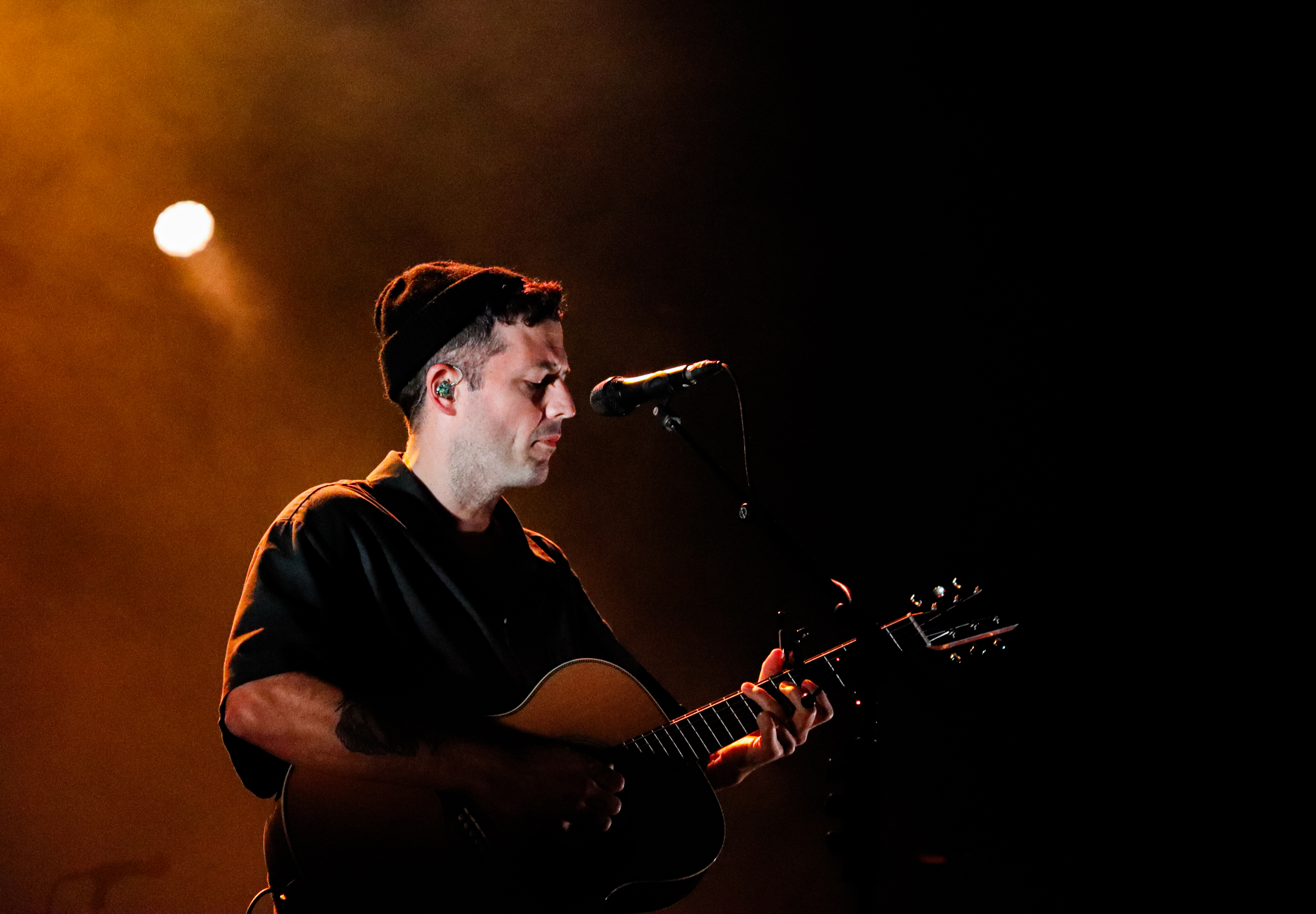 A musician performing on stage, playing an acoustic guitar while singing into a microphone, with warm lighting creating an atmospheric backdrop.