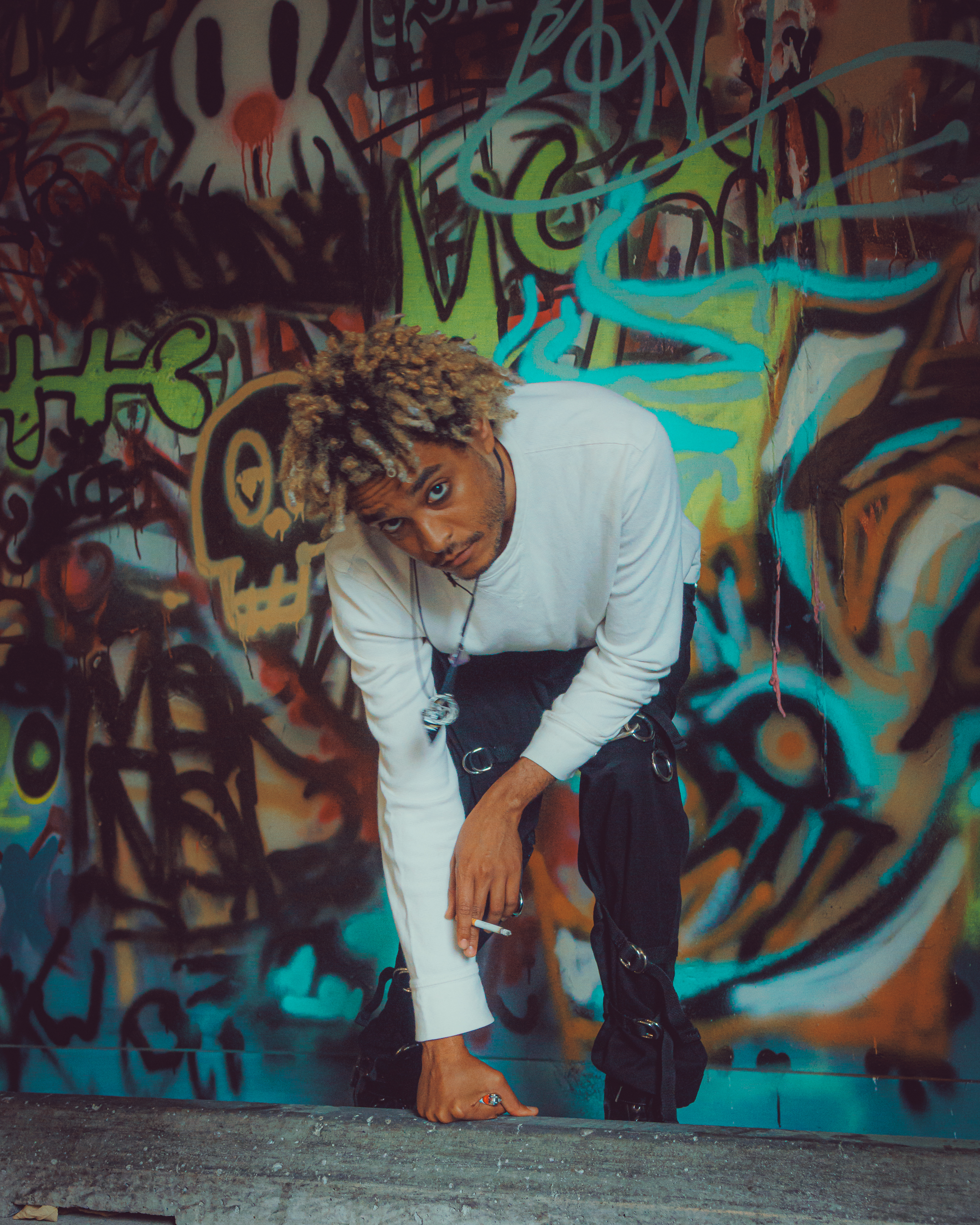A person with curly blonde hair kneeling on the ground, wearing a white long-sleeve shirt and black pants with chains, holding a cigarette, in front of a colorful graffiti-covered wall.