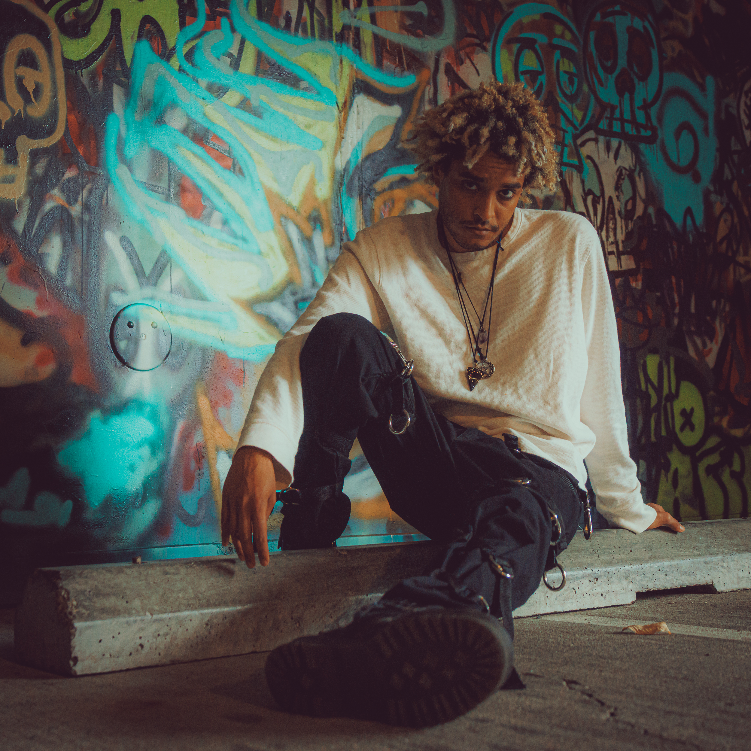 A person sitting against a colorful graffiti wall, wearing a white long-sleeve shirt and black pants, with accessories and a contemplative expression.